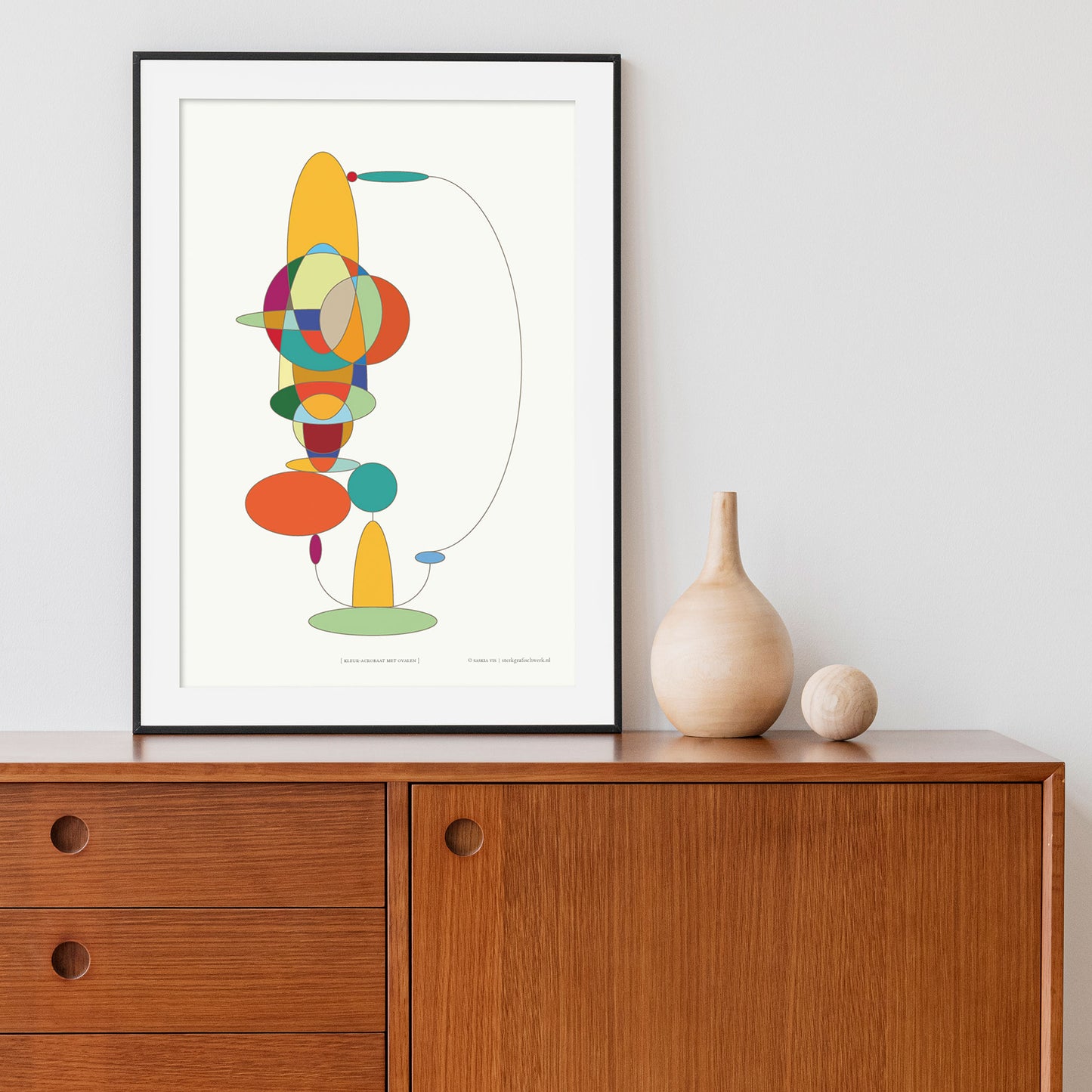Productafbeelding, poster "Kleur-acrobaat met ovalen", staande op een wandmeubel in een zwarte lijst tegen een wittte wand