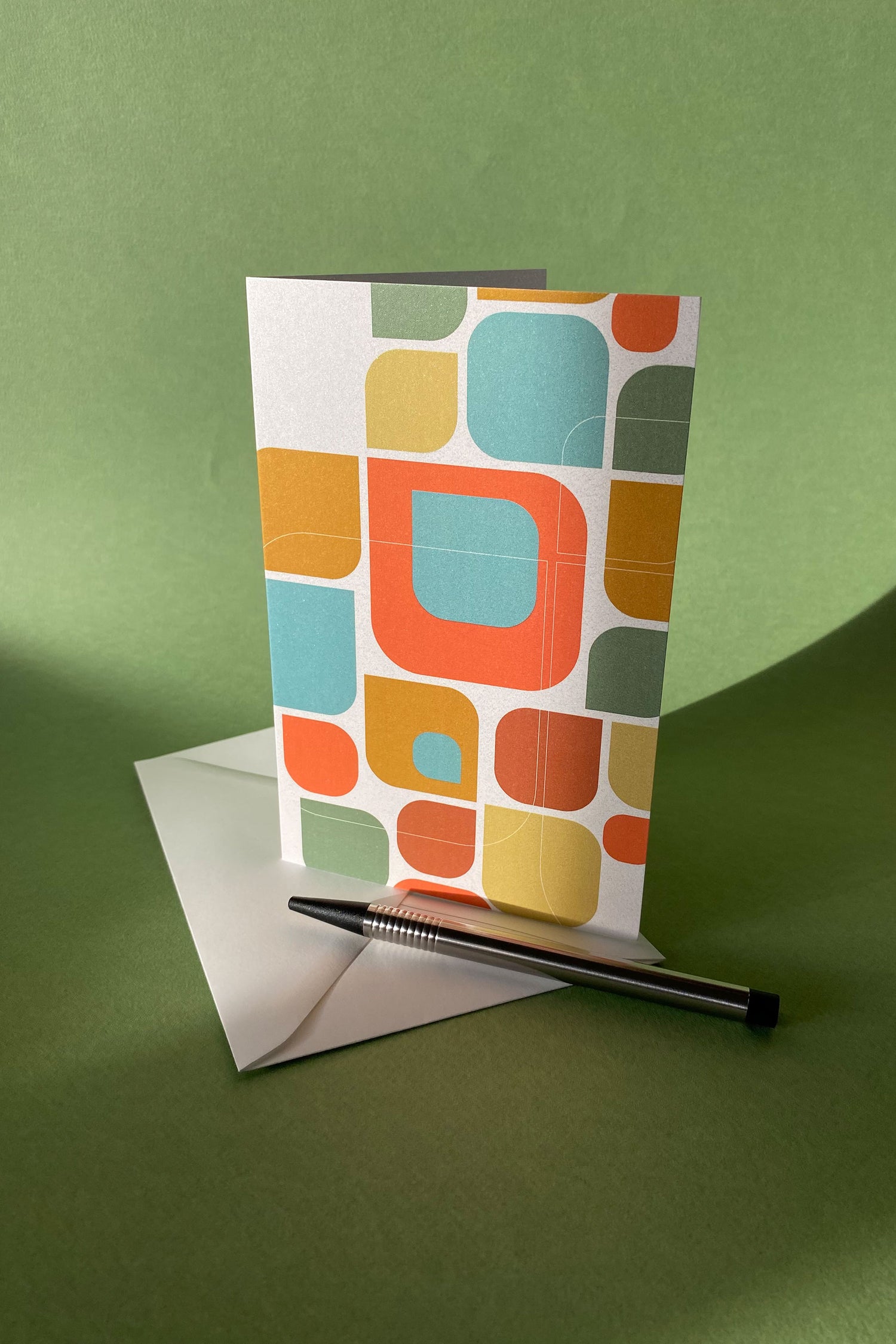 Sfeerfoto van product wenskaart "Kleurmotief op wit" met een envelop en een pen tegen een groene achtergrond
