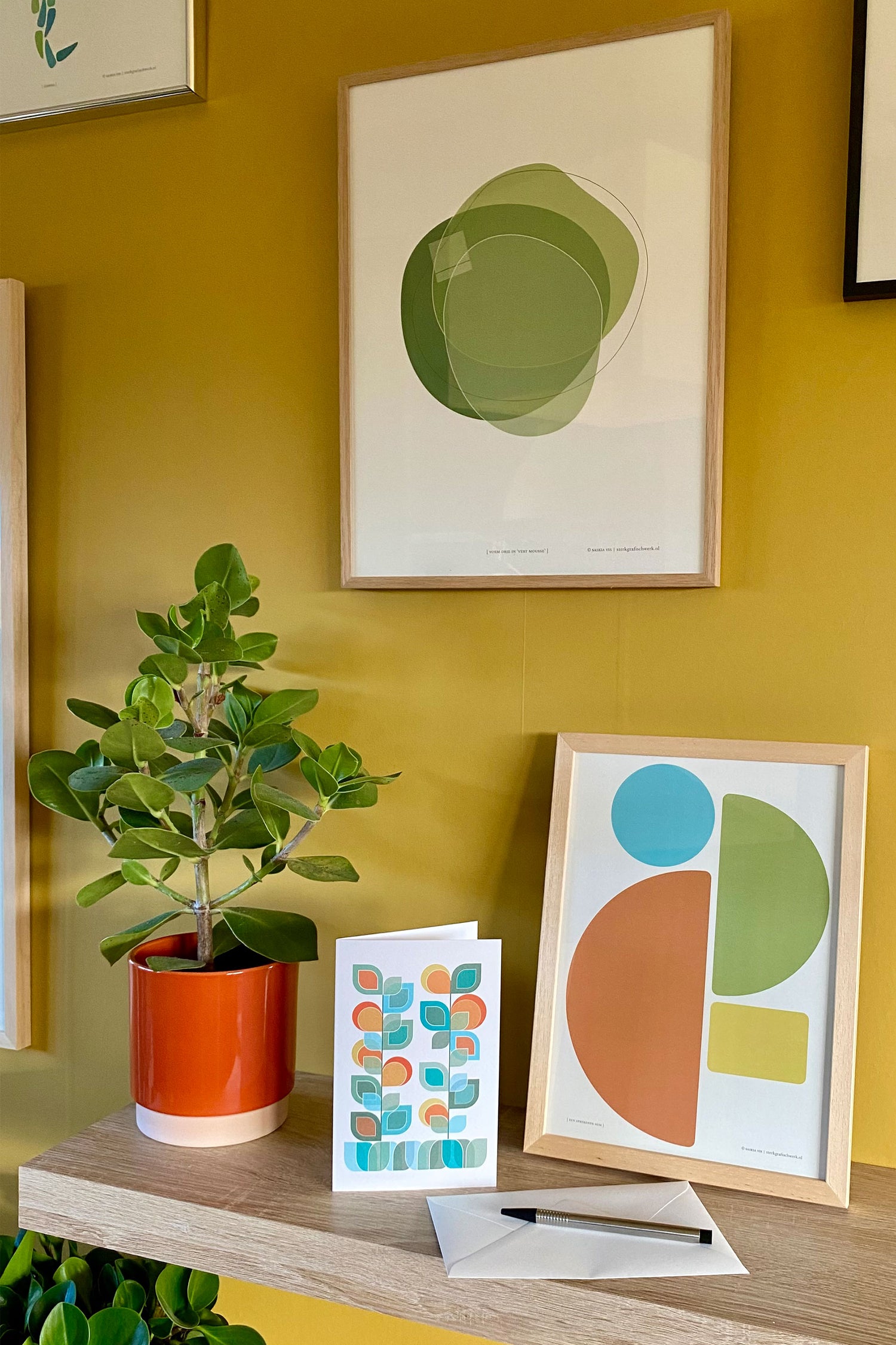 Sfeerfoto met drie producten, poster "Vorm drie in vert mousse" samen met "Een sprekende som" en wenskaart "Vaasje Bloemen 4" en een plant